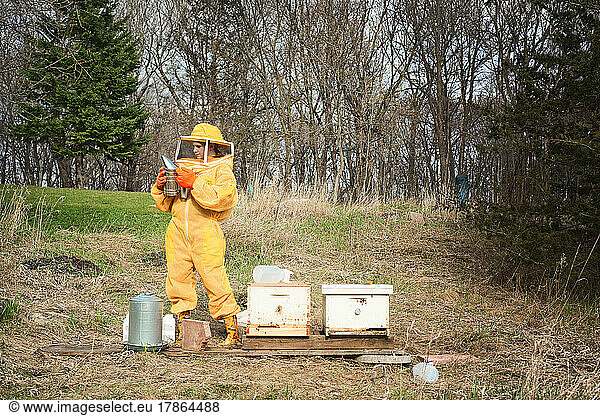 Girl in beekeeping suit standing by two hives with a smoker.