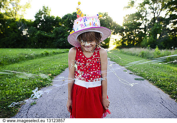 Girl in birthday cake hat standing on road while party strings sprayed on her