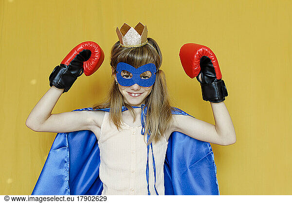 Girl in cape wearing mask and boxing gloves against yellow background