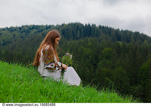 girl in embroidery with a bouquet of wild flowers sits on green grass in the carpathian mountains