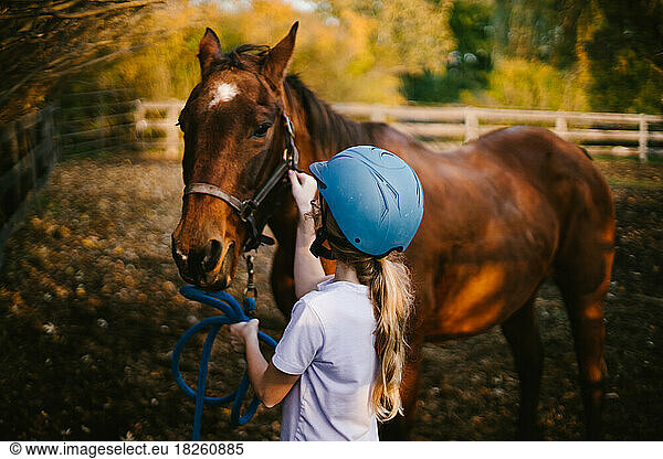 Girl in riding helmet unhooks bridle from brown horse in fall