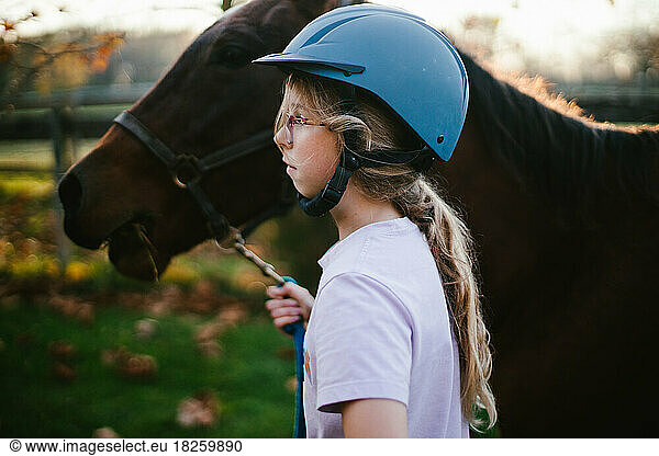 Girl in riding helmet walks horse through stalls in sunlight