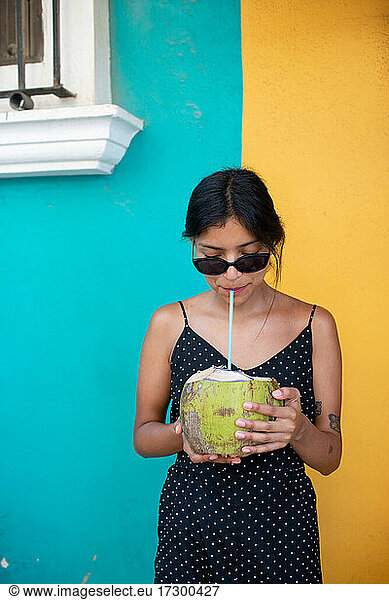 Girl in sunglasses drinking coconut in front of colorful wall