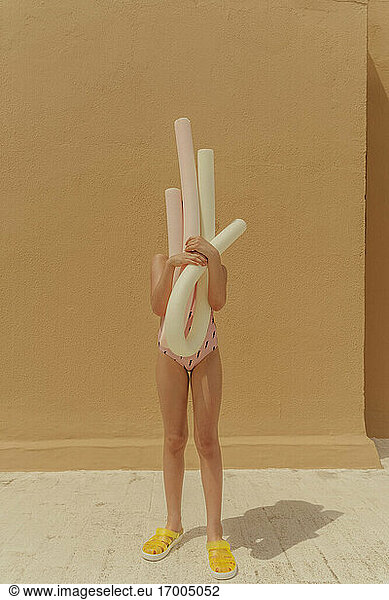 Girl in swimsuit hiding her face behind pool noodles