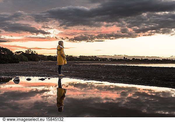 Girl in yellow coat standing on beach at sunset with reflection