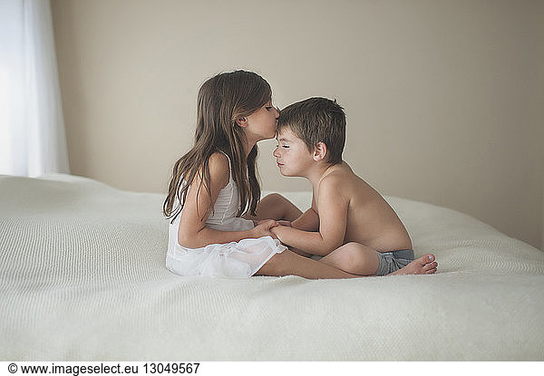 Girl kissing brother on forehead while sitting on bed at home