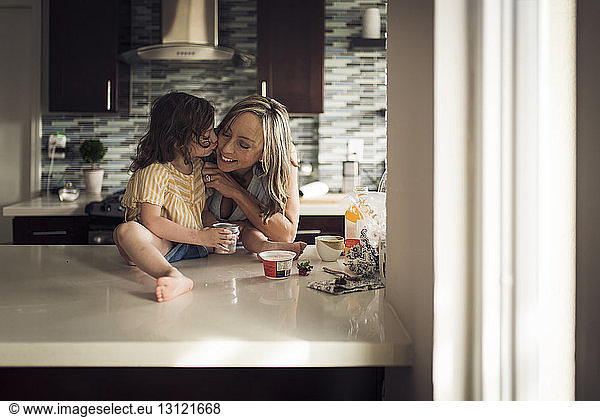 Girl kissing mother sitting on kitchen counter at home