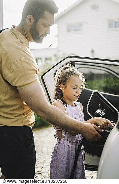 Girl learning to charge electric car while standing by father
