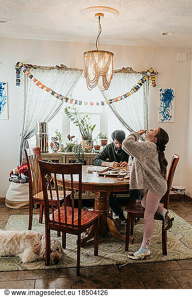 Girl licks finger while building gingerbread house with dad