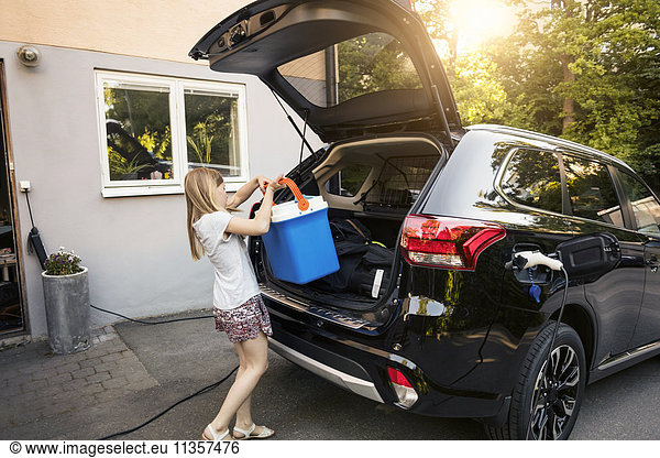 Girl loading cooler in black car trunk against house