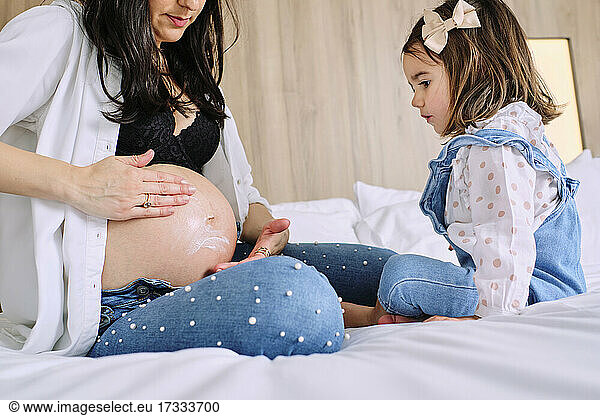 Girl looking at mother applying moisturizer on belly while sitting on bed
