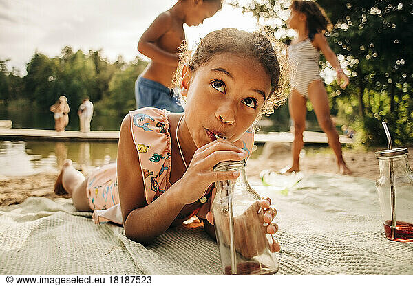 Girl looking away while drinking juice from bottle lying down on picnic blanket during vacation