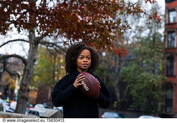 Girl looking away while holding American football in city