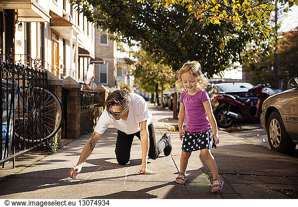 Girl looking down while father drawing hopscotch on footpath