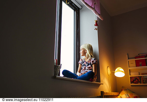 Girl looking through window while sitting on sill
