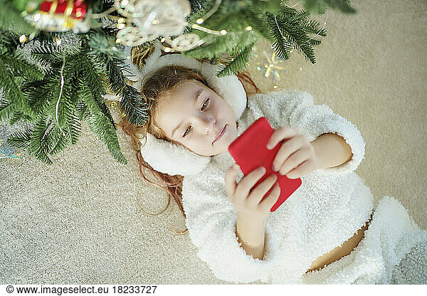 Girl lying on carpet by Christmas tree using smart phone at home