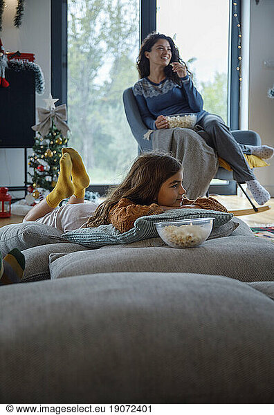 Girl lying on pillows by mother having popcorn at home