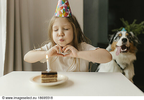Girl making heart gesture and blowing candle on cake at home