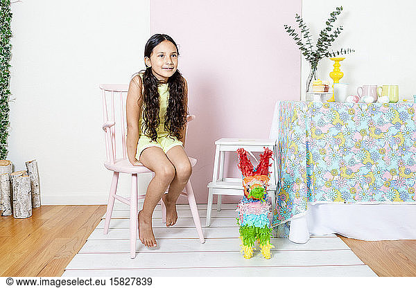 Girl on overall sitting in pink chair at home party decor by pinata