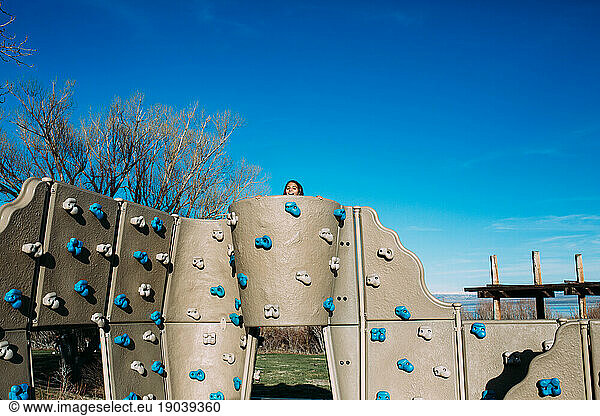 Girl peeks over a rock wall at a playground on a sunny blue sky day