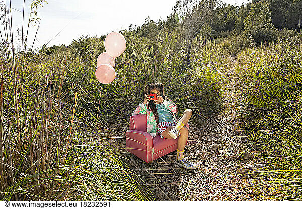 Girl photographing through toy camera sitting on pink armchair in field