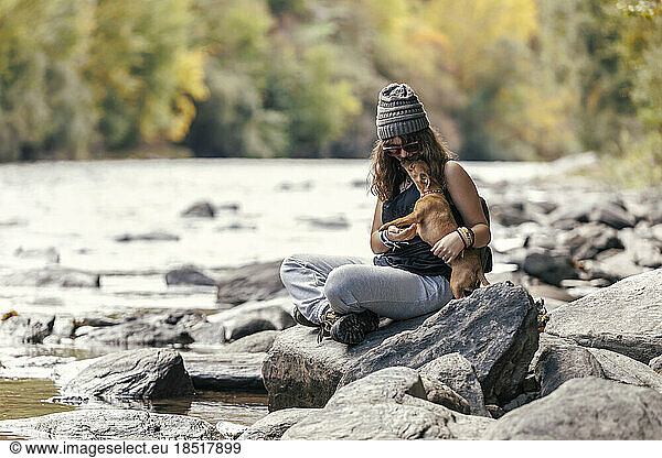 Girl playing with dog sitting cross-legged on rock near river