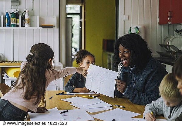 Girl pointing at teacher showing drawing during art class in day care center