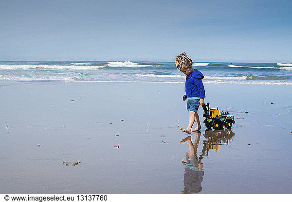 Girl pulling toy truck while walking at beach against cloudy sky