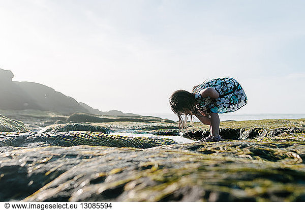 Girl searching something while bending over rocks against clear sky
