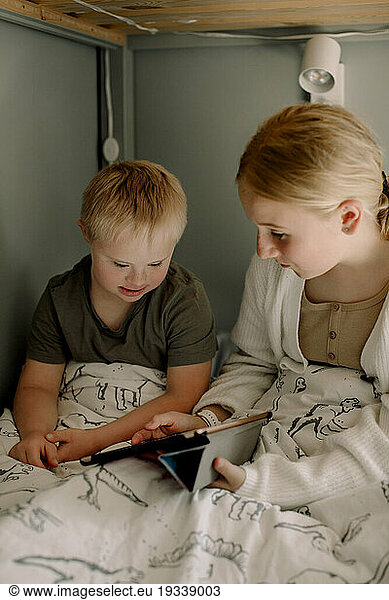 Girl sharing digital tablet with brother sitting on bed at home