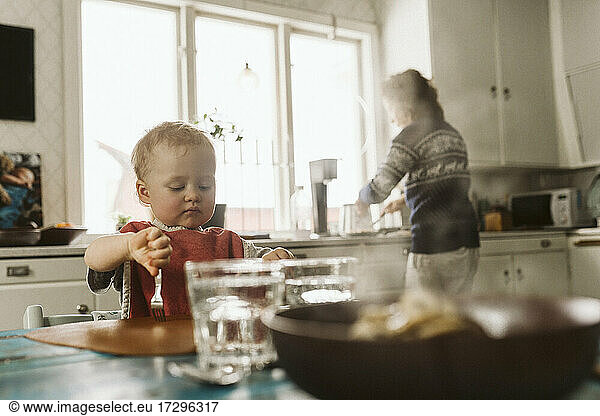 Girl sitting at dining table while mother prepares food in kitchen