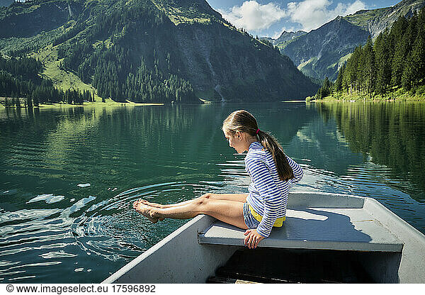 Girl sitting on bow of rowboat dipping feet in lake