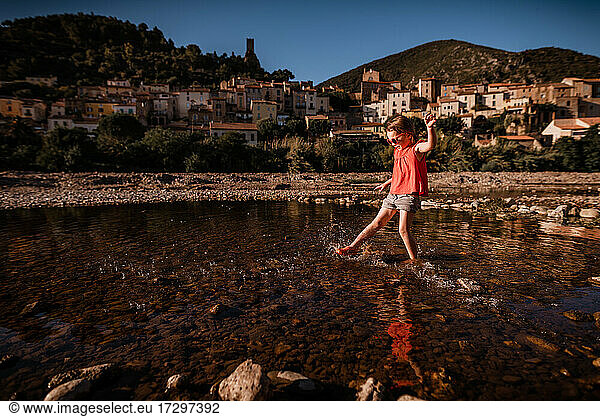 Girl splashes water in river in France with village in background