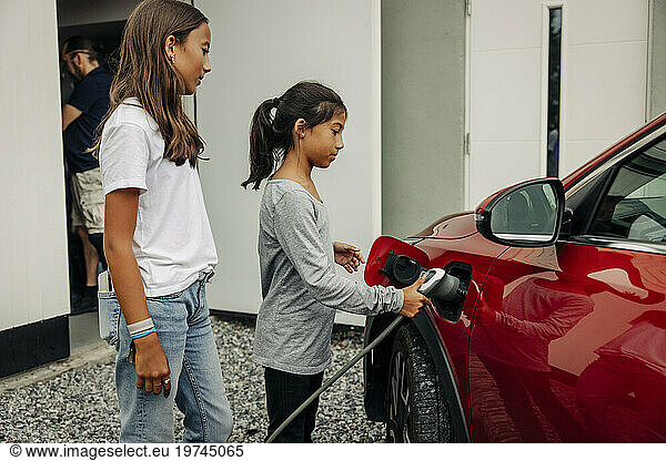 Girl standing next to sister charging electric car near house