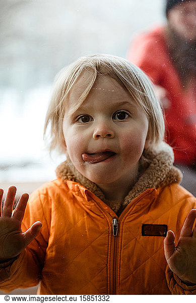girl toddler pushes on window looks through glass with twisted tongue