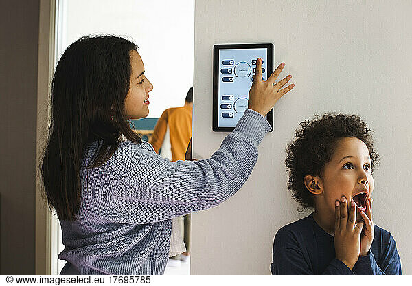Girl using home automation device while standing with surprised brother against wall at home