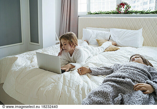 Girl using laptop lying on bed by brother at home