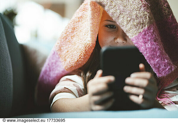 Girl using smart phone lying under blanket at home