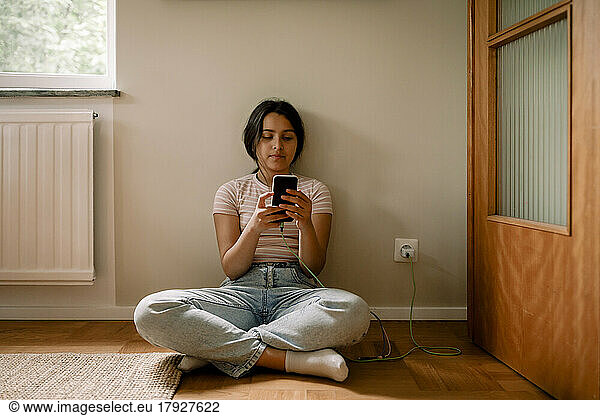 Girl using smart phone while sitting cross-legged on floor against wall at home