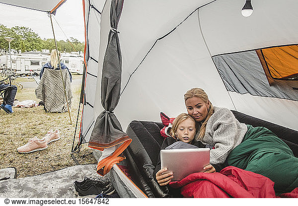 Girl watching digital tablet while lying with mother in tent at campsite