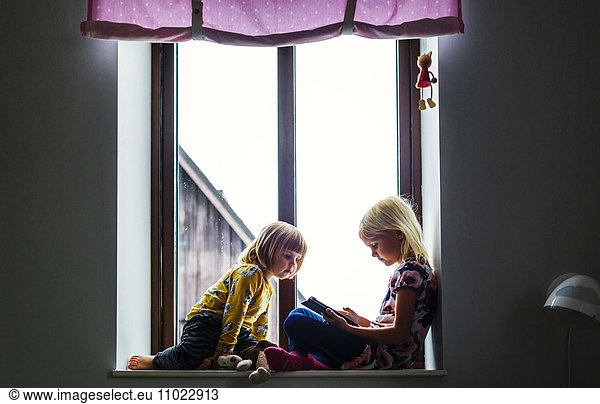 Girl watching sister using digital tablet on window sill