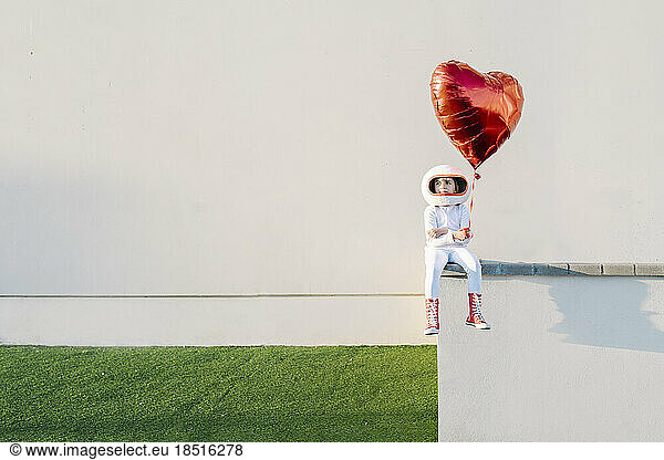 Girl wearing astronaut costume sitting on wall holding heart shape balloon