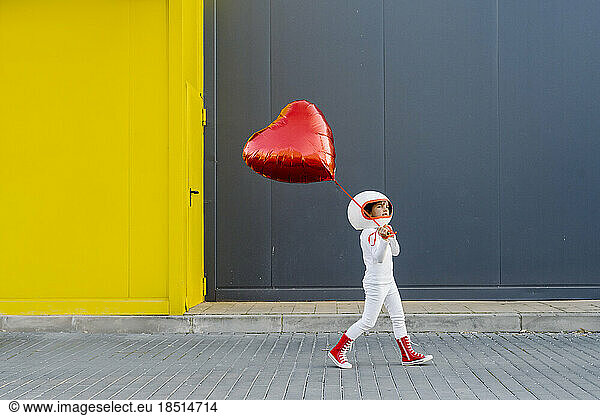 Girl wearing astronaut costume walking with heart shape balloon near yellow wall