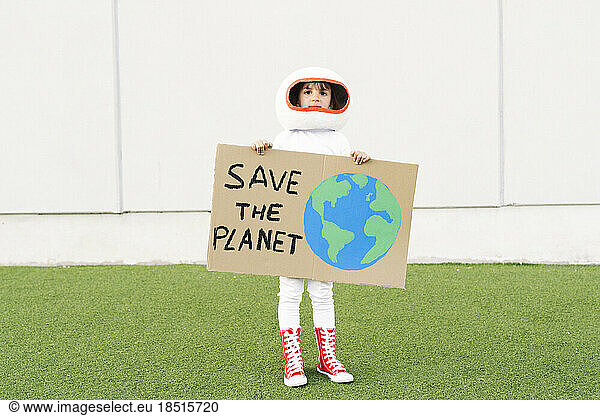 Girl wearing astronaut dress holding Save The Planet placard standing in front of wall