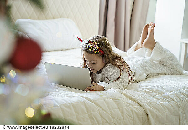 Girl wearing Christmas headband lying on bed and using laptop at home