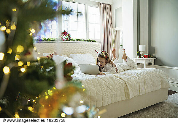 Girl wearing Christmas headband using laptop on bed at home