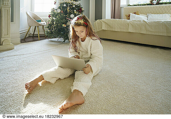 Girl wearing Christmas headband using laptop sitting on carpet at home