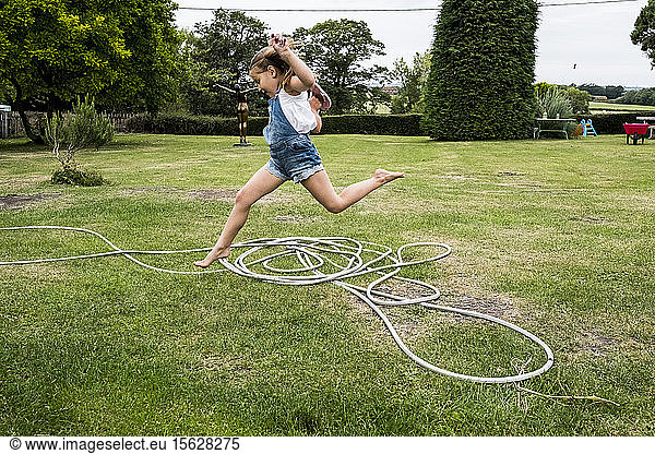 Girl wearing denim dungarees jumping over a garden hose on a lawn.
