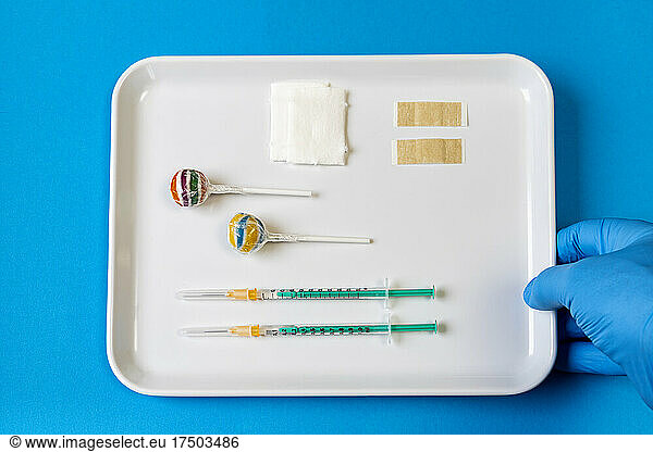 Girl wearing glove holding tray of lollipops and vaccine injections on blue background