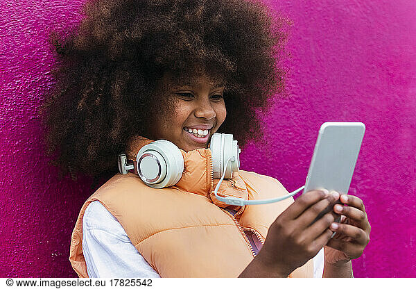 Girl wearing headphones using mobile phone leaning on pink wall